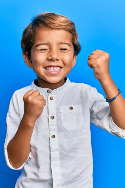 The image features a young boy with a joyful expression, celebrating enthusiastically. He has short, light brown hair and is wearing a light grey button-up shirt with a chest pocket. His eyes are cheerfully closed, and his mouth is open in a bright smile, showing his teeth. Both of his fists are clenched in a victorious gesture. He is also wearing a simple blue bracelet on his wrist. The background is a vivid blue, which beautifully contrasts with his light-colored shirt and highlights his exuberant pose.