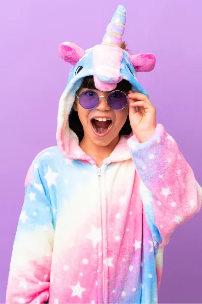 This image features a person wearing a vibrant, pastel-colored unicorn onesie with a hood that has a unicorn face, ears, and a horn. The onesie is a delightful mix of blue, pink, and purple with star patterns. The person is also wearing round, purple-tinted sunglasses and is lifting the glasses with one hand, playfully revealing their eyes. They are smiling broadly and appear to be having a great time. The background is a plain, light purple, complementing the colors of the onesie.